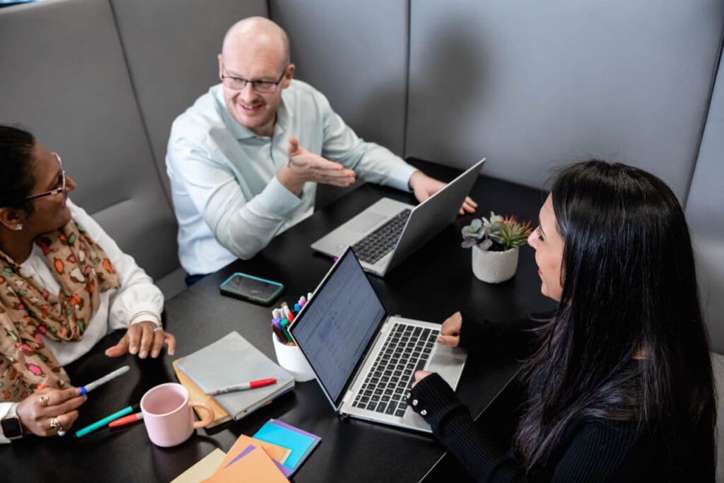 A group of Ultima employees sat a working space together with laptops, pens and notebooks