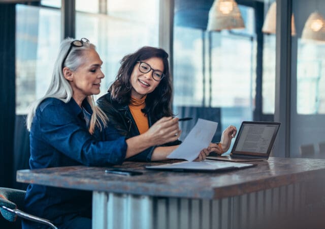 Two older women look at charts on paper with a laptop besides them