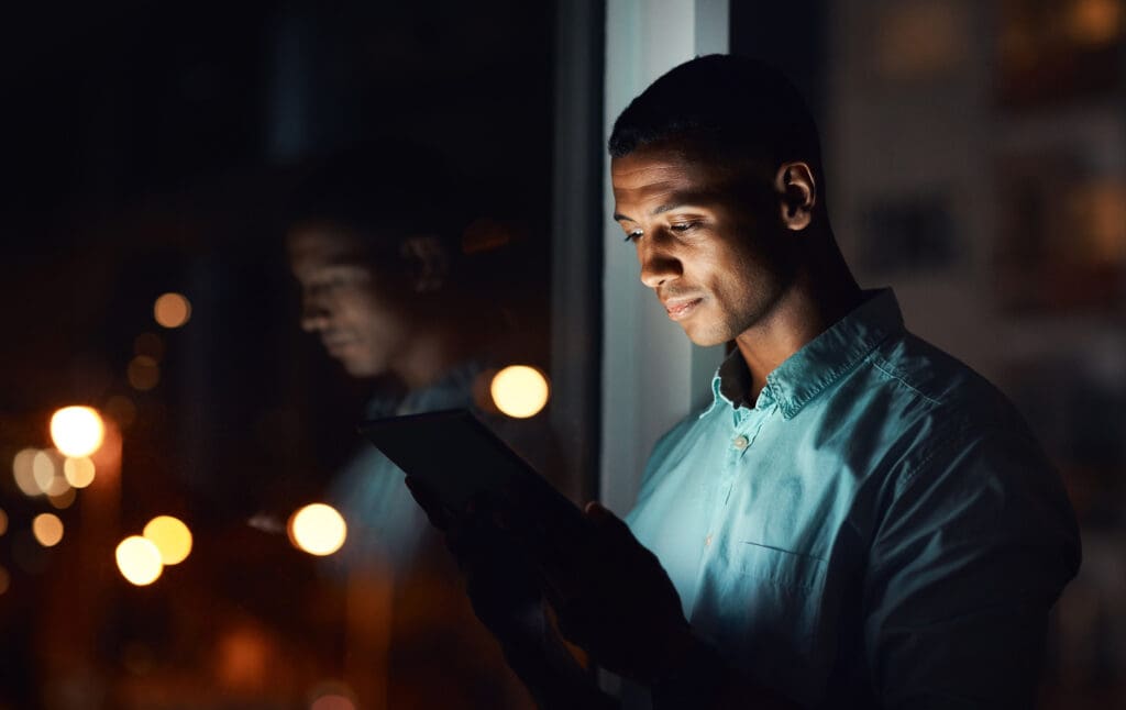 A man stands by a window in a dark room looking at a tablet screen