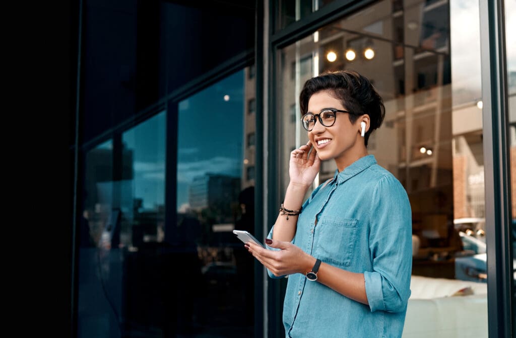 A young women stands outside an office building holding her phone and wearing headphones
