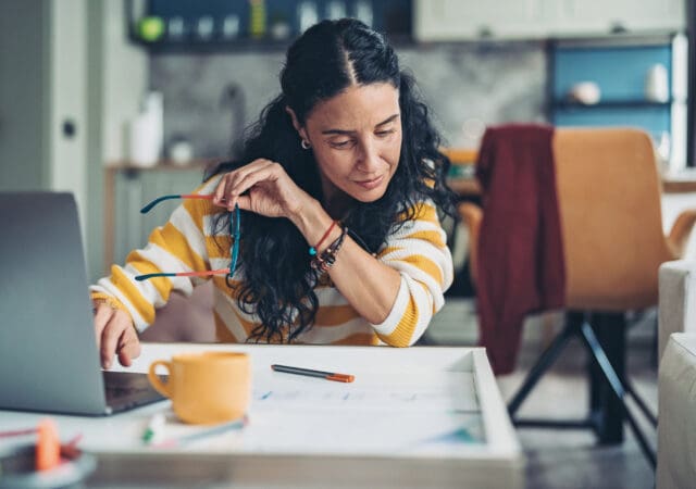 A women looks at her notes while holding her glasses and looking at a laptop screen