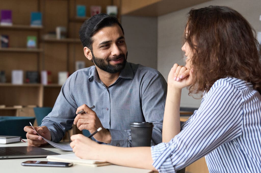 A man and a woman have a conversation at a desk while holding notebooks and pens