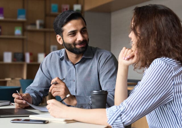 A man and a woman have a conversation at a desk while holding notebooks and pens