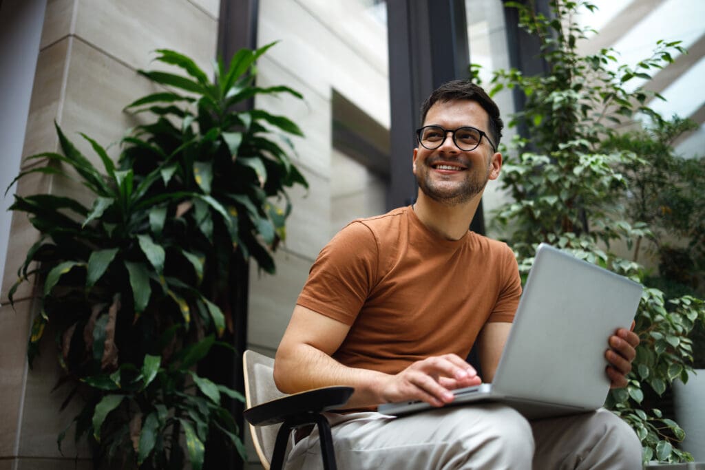 A man smiles while working on a laptop in a verdant workplace
