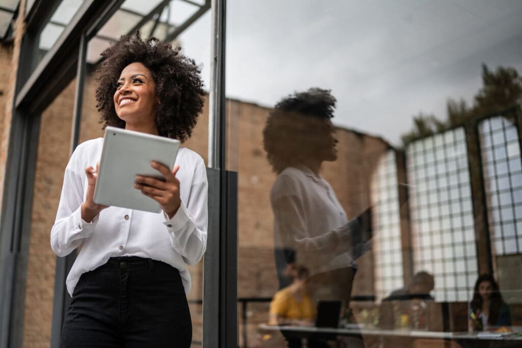 A women smiles as she looks away while holding a tablet in front of a modern office building
