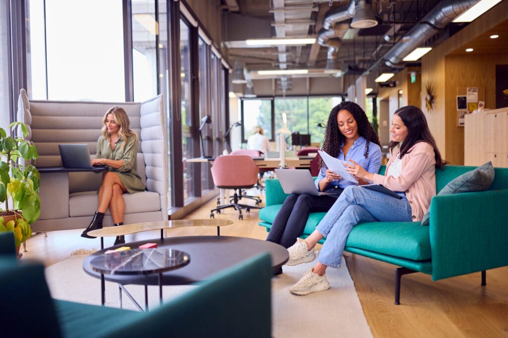 Two women look at a document while another works on a laptop in a modern, comfortable workplace