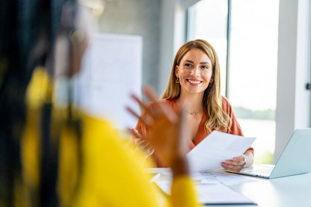 A woman smiles as she has a conversation with another woman sitting round a table in a bright workplace