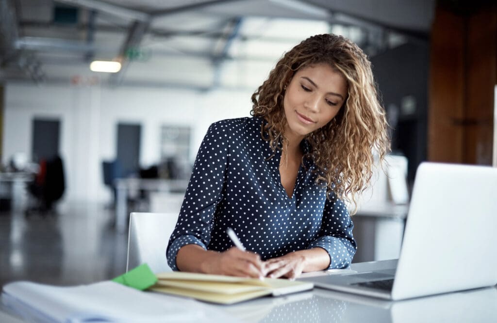 A young women writes in a notebook with a laptop in front of her
