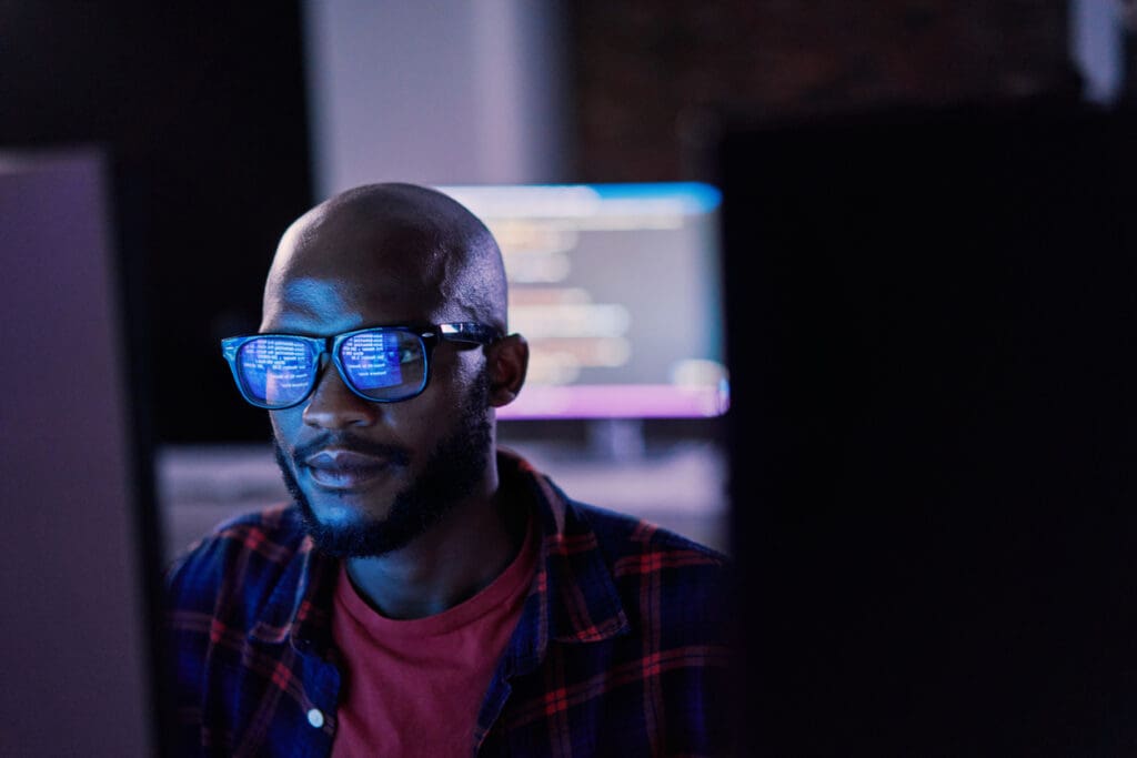 Person wearing glasses with blue screen reflection, sitting at computer in dimly lit room