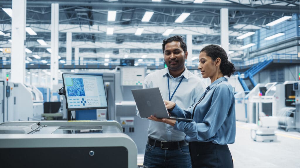 Two professionals (male and female) in a large, well-lit modern space, surrounded by computers; the female is holding a laptop and pointing to show the male something on screen.