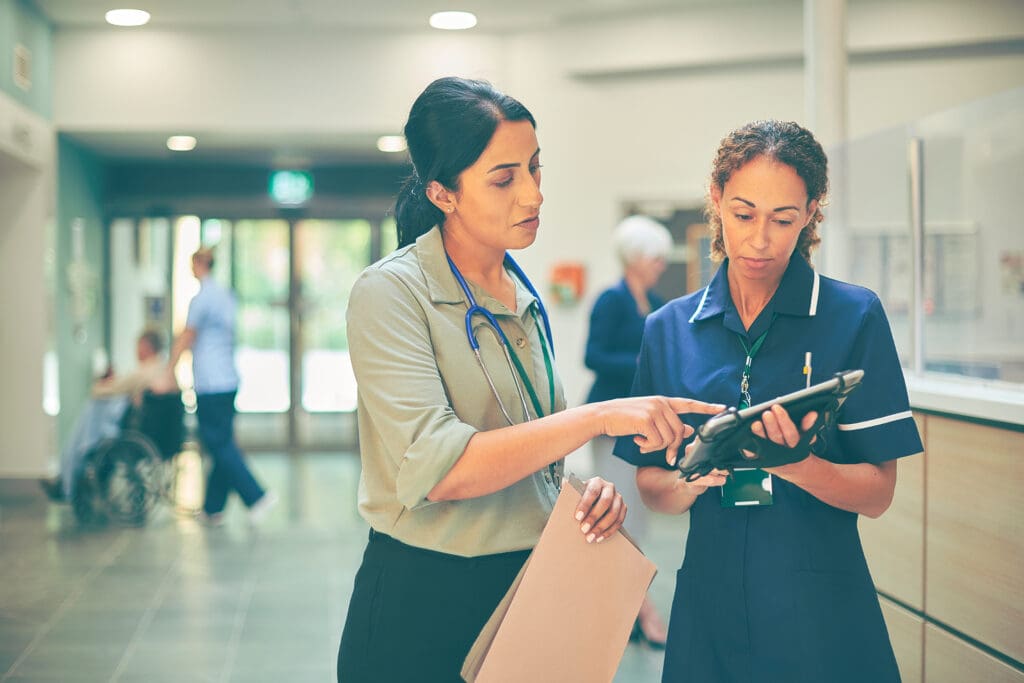 Two healthcare professionals reviewing patient information on a digital tablet in a hospital reception area, highlighting collaborative clinical decision‑making and modern NHS digital workflows